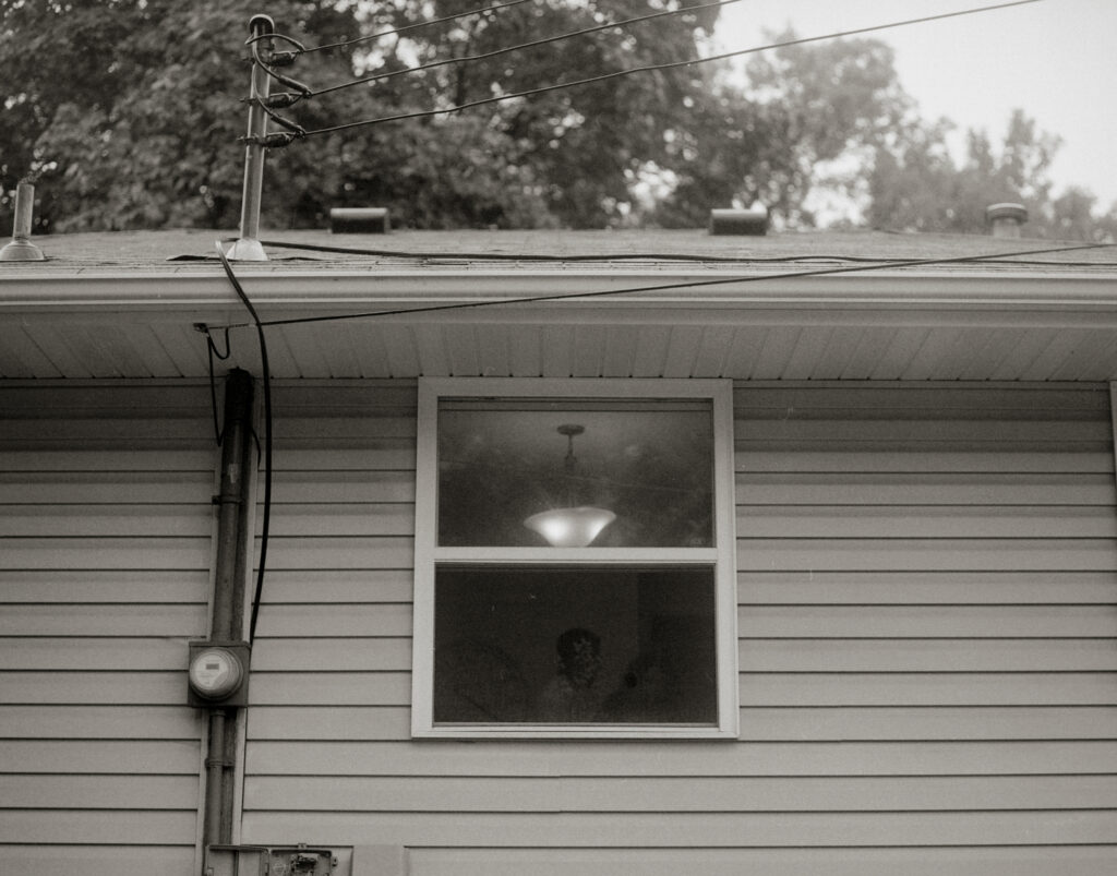 Side of house with beige siding and electric meter, with light fixture turned on inside the window.