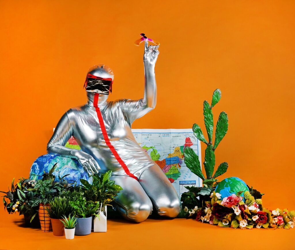 Person wears silver full-body jumpsuit with red stripe and AR goggles, surrounded by plants, a map, and a globe, with orange background.