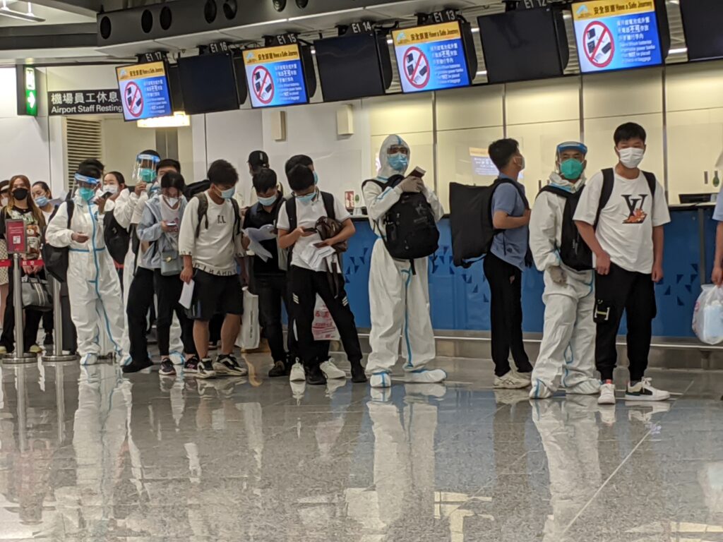 People standing in line at airport, wearing surgical masks and hazmat suits.