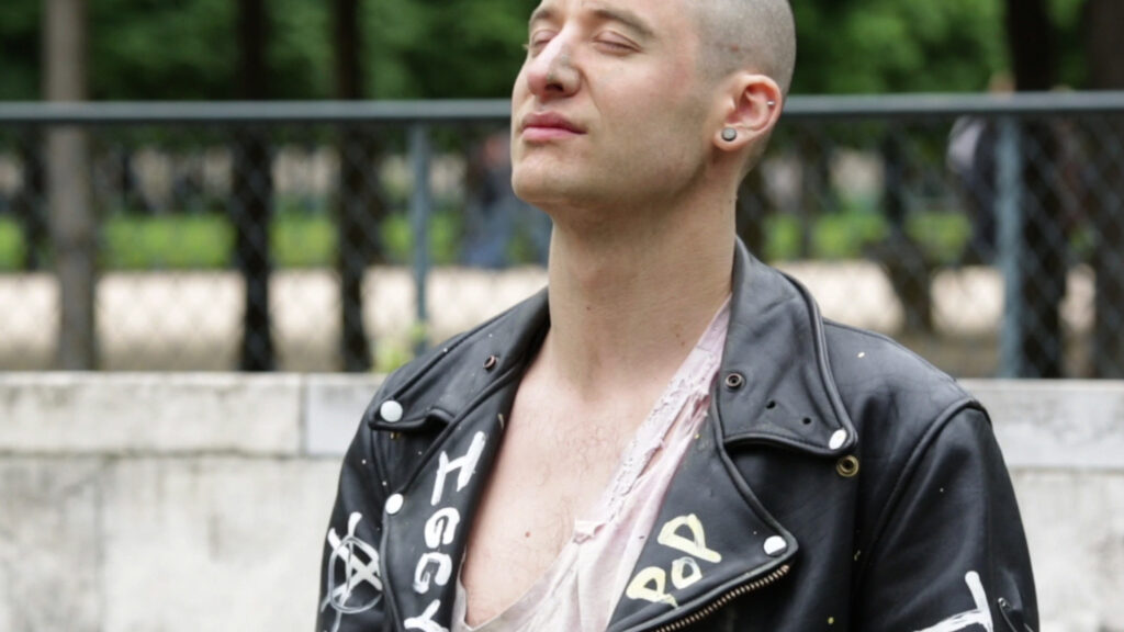 Light skinned person with a shaved head and leather jacket tilts face upwards with closed eyes