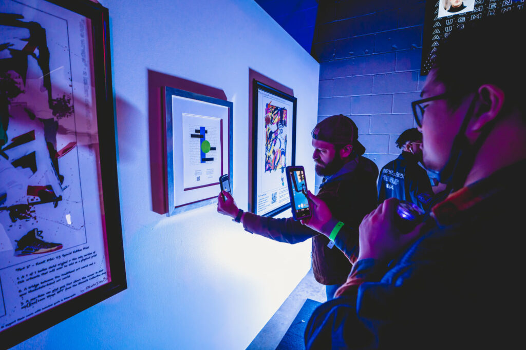 People hold smartphones up to artwork framed on walls, in blue light.