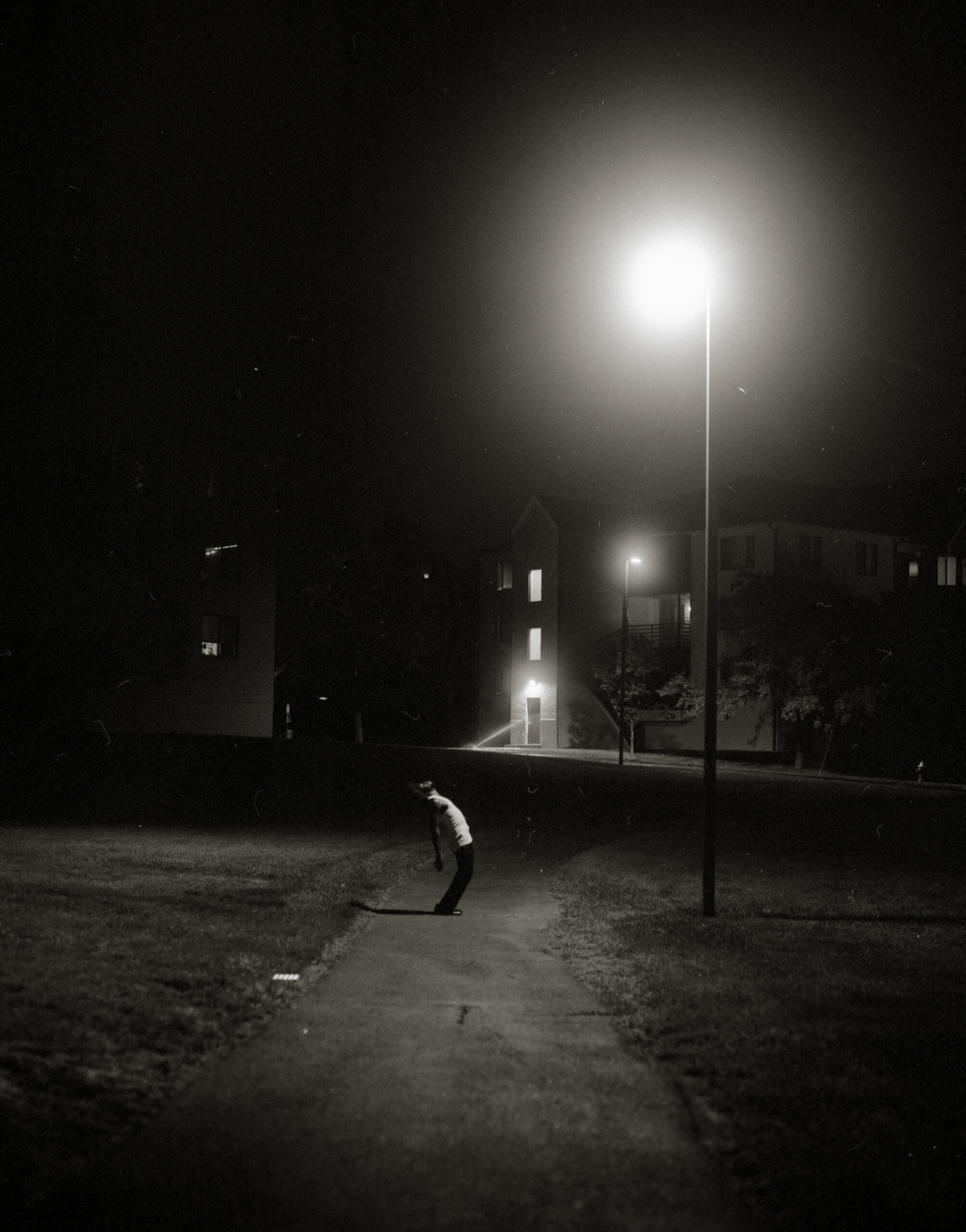 Person stands on dirt path and leans back underneath tall street lamp at night.