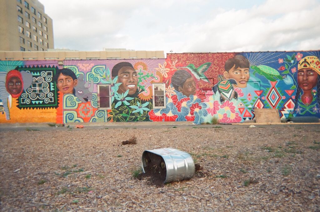 Colorful mural with faces of diverse people, behind mulch-covered lot.