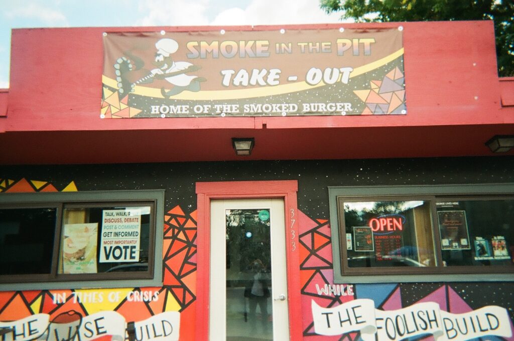 Red facade of restaurant with signs in windows and sign reading "Smoke in the Pit".