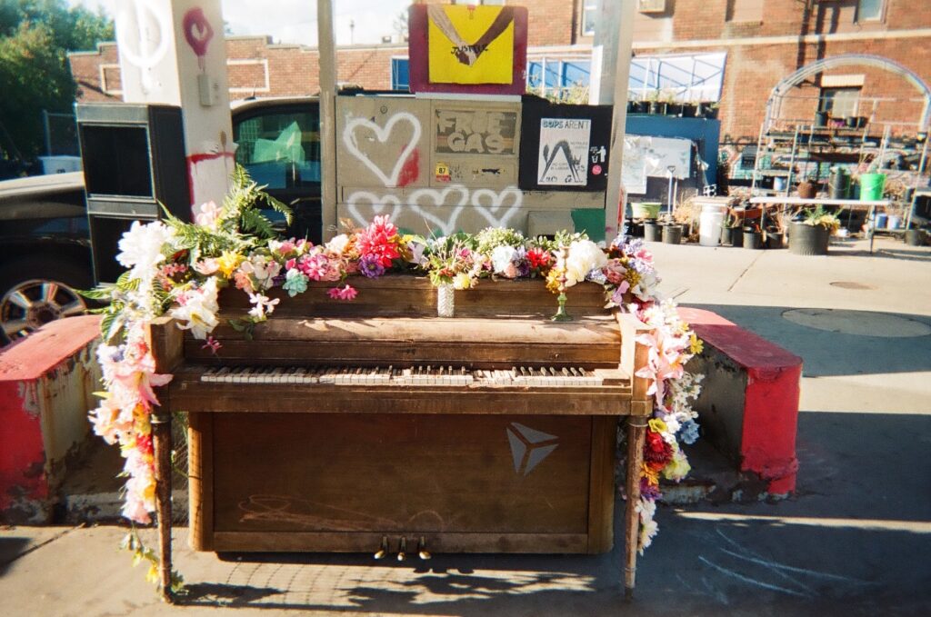 Floral wreath covers upright piano, at out-of-use gas station with many painted signs.