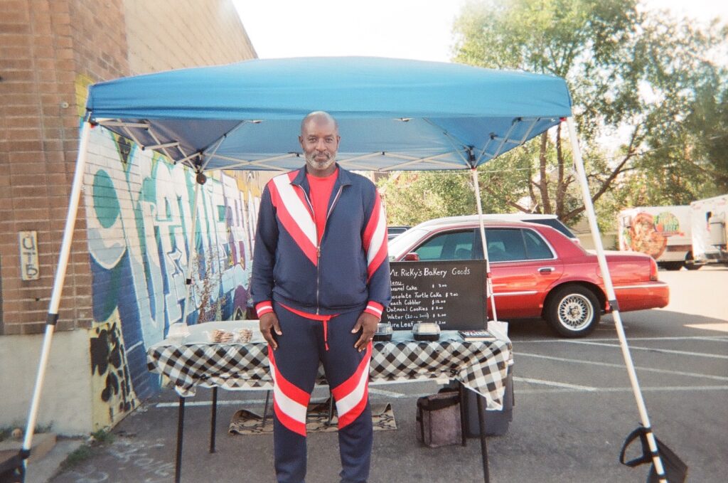 Person with dark skin wears striped tracksuit and stands in front of a table with blue awning.