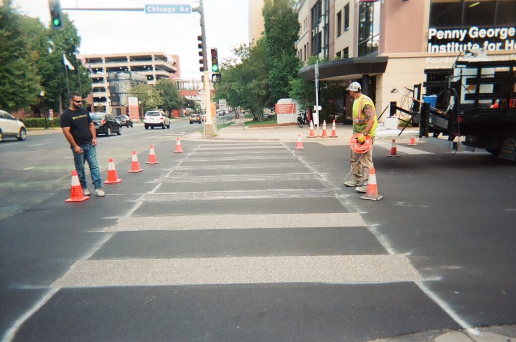 Construction worker places orange cones in crosswalk, with a pedestrian standing opposite.