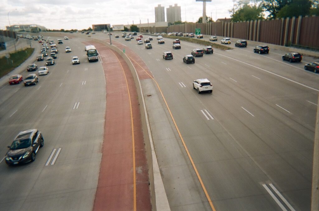 Many cars drive on freeway, with median in center and high rise building sin background.