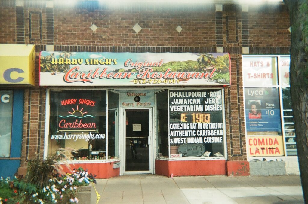 Restaurant facade with neon signs in window, and sign reading: Harry Singh's Original Caribbean Restaurant.