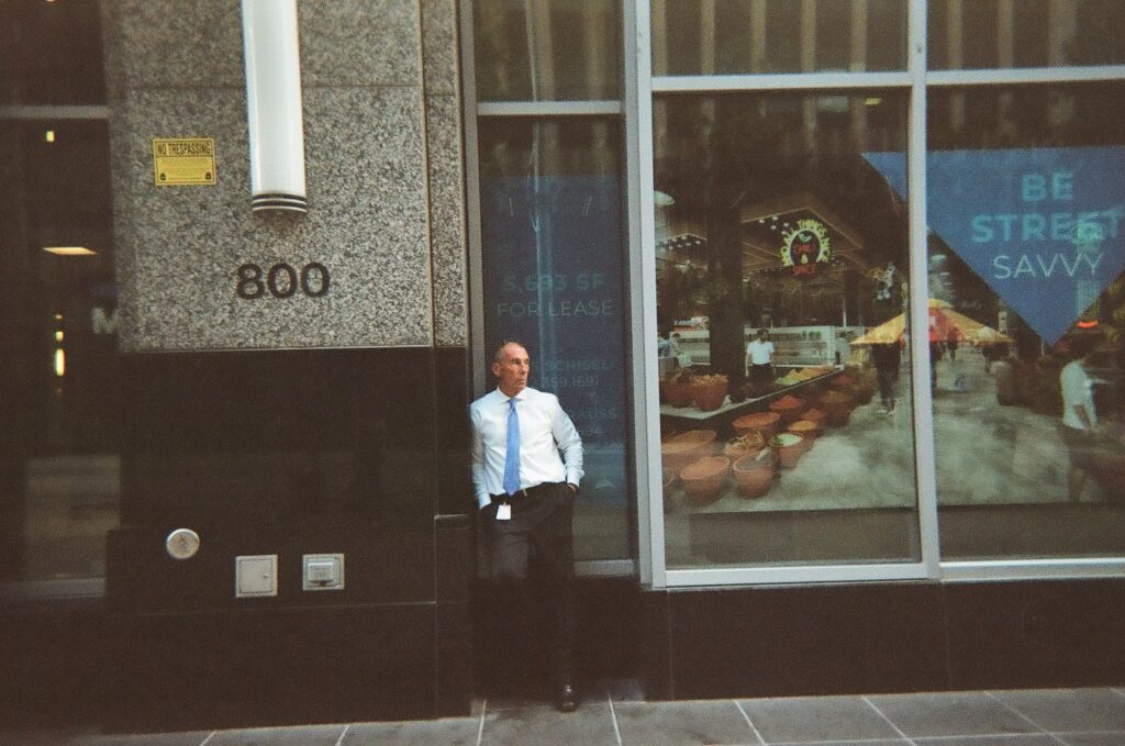 Person with light skin, black slacks, white dress shirt, and blue tie leans against wall of black and gray marble building.