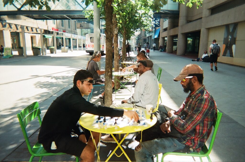 Many people play chess on outdoor tables on city sidewalk.