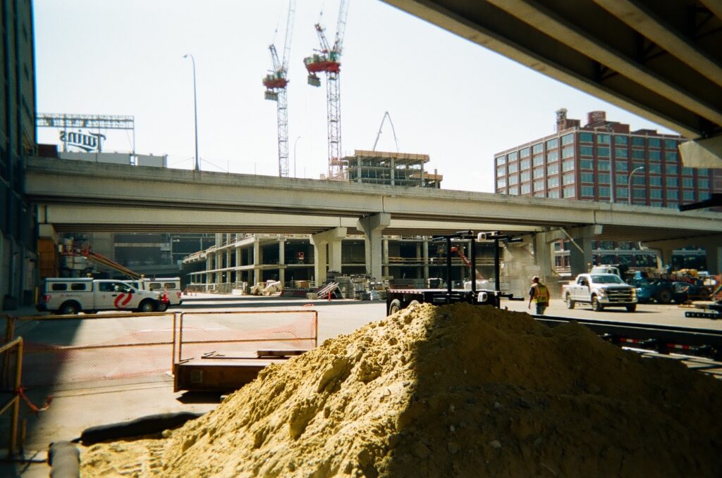 Construction zone with dirt pile in foreground, overpass in mid-ground, and building, cranes, and Twin stadium sign in background.