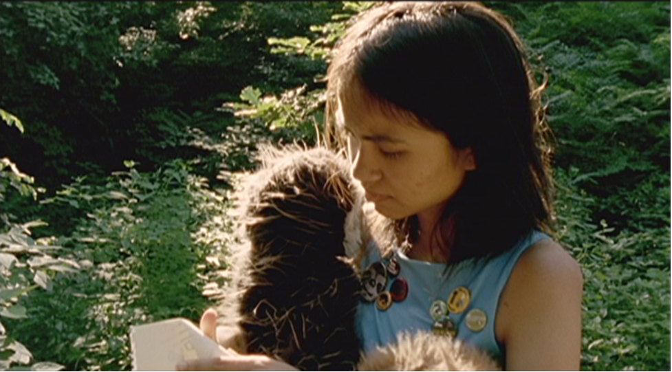 Child with shoulder-length brown hair holds stuffed animal, with sunlight on face and greenery behind.