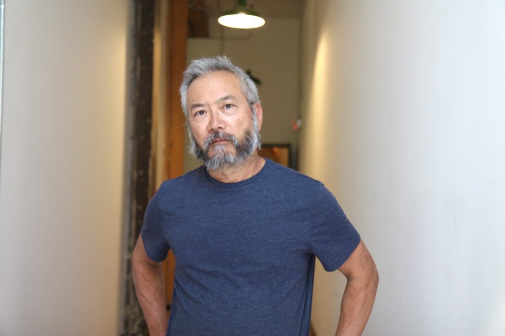 Asian person with short salt-and-pepper hair and beard wears blue t-shirt and looks at camera with hands on hips.