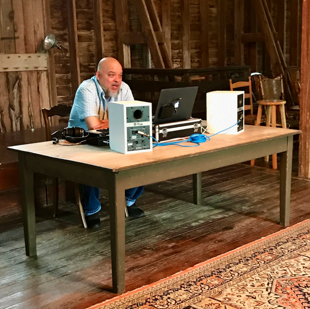 Person sits at large table in barn, with speakers and laptop.