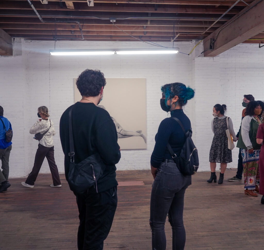 Two people stand facing artwork in a gallery, with others mingling in the background.