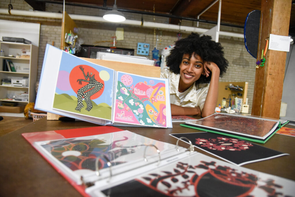 Dark skinned person smiles at a table with colorful artwork displayed in binders.
