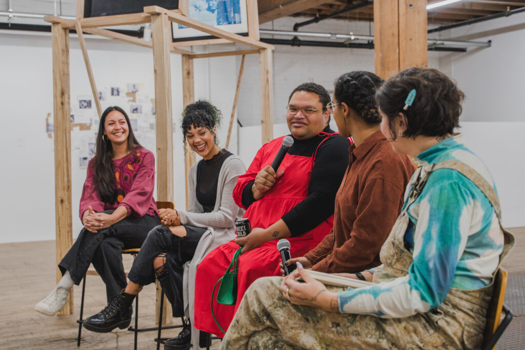 5 people sit on chairs in a studio gallery, one holding a microphone.