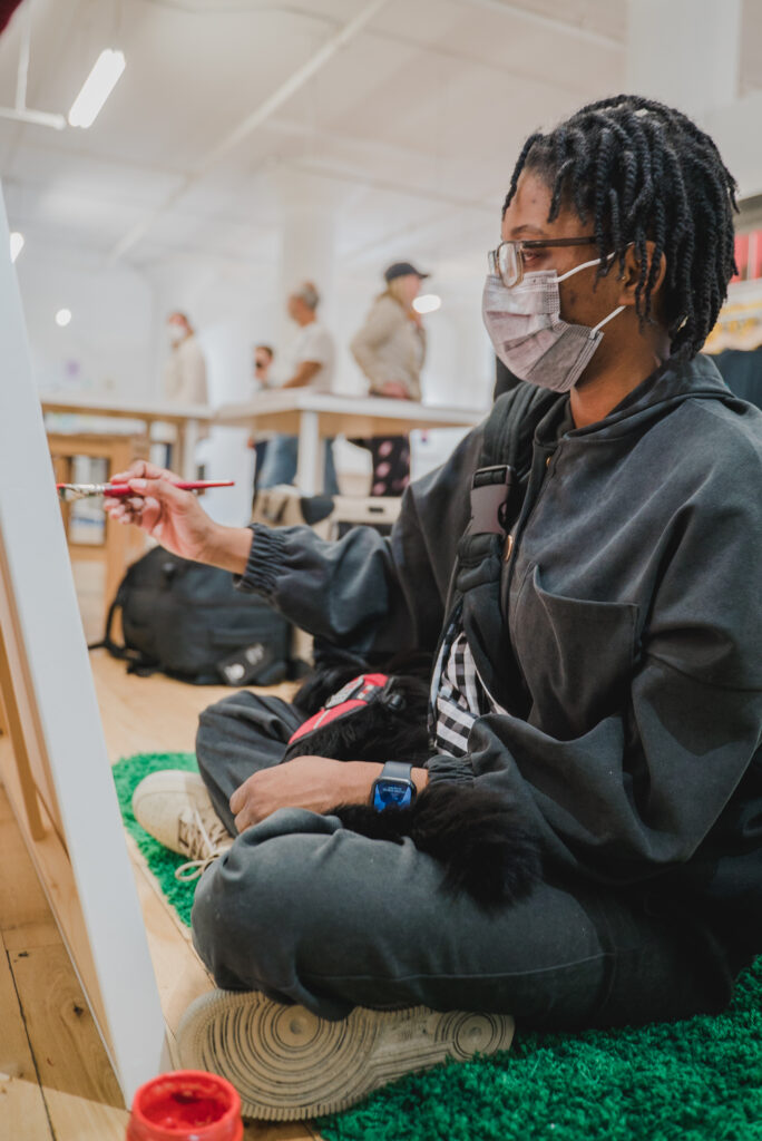 Masked dark-skinned person sits in front of a canvas holding a paintbrush.