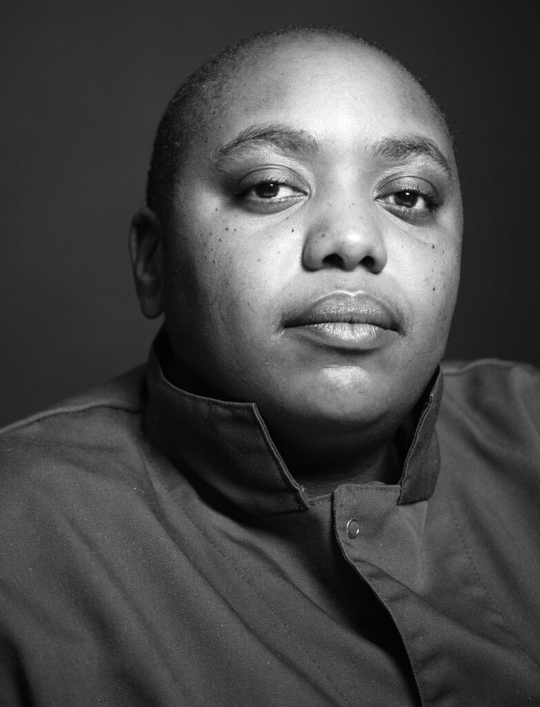 Black and white portrait of person with medium dark skin, shaved head, and collared shirt looking directly at the camera.