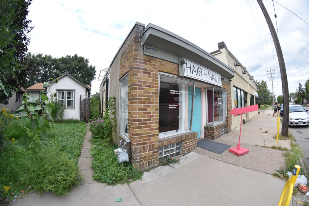 Fisheye lens photo of gallery with brick facade and teal door, and white house and green lawn beside it.