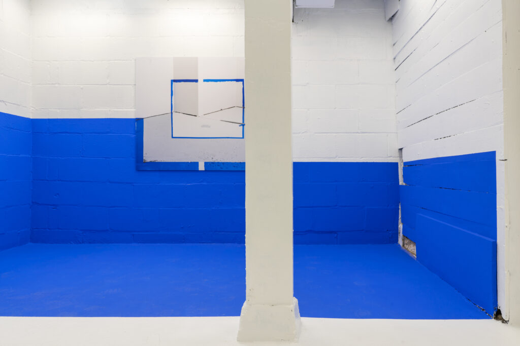 Gallery with bright blue paint on floor and lower half of wall, with white column and upper wall, and rectangular 2D artwork on wall.