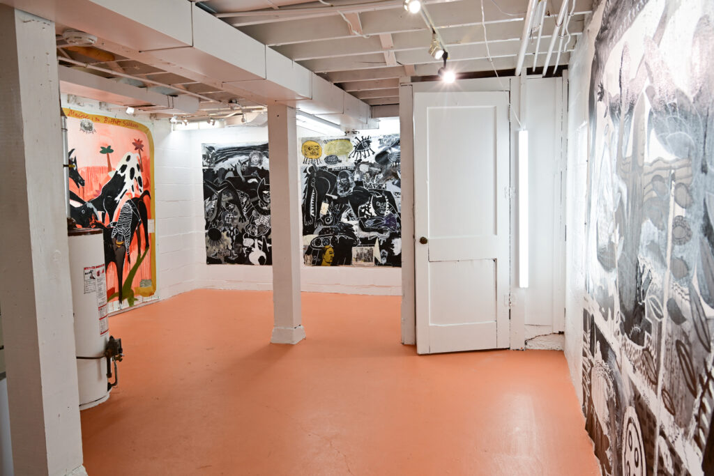 Basement gallery with peach floor, orange and dark gray paintings on walls, and white column and door.