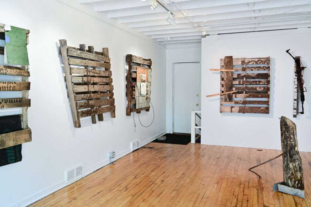 Gallery with white walls and wood floor, with artworks on walls made from wooden pallets.