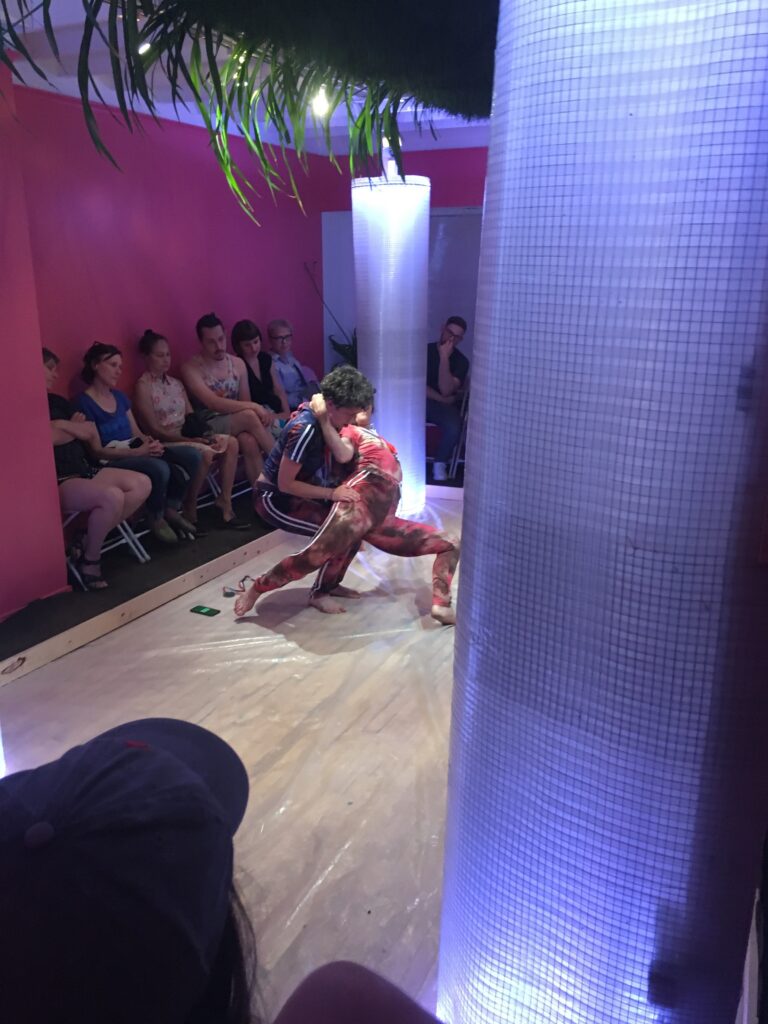 Two dancers kneel in contact in small gallery, with row of audience behind them.