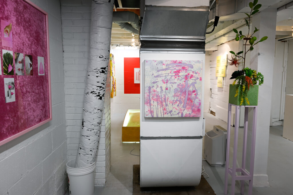 Basement gallery with pink and red paintings hung on white walls.