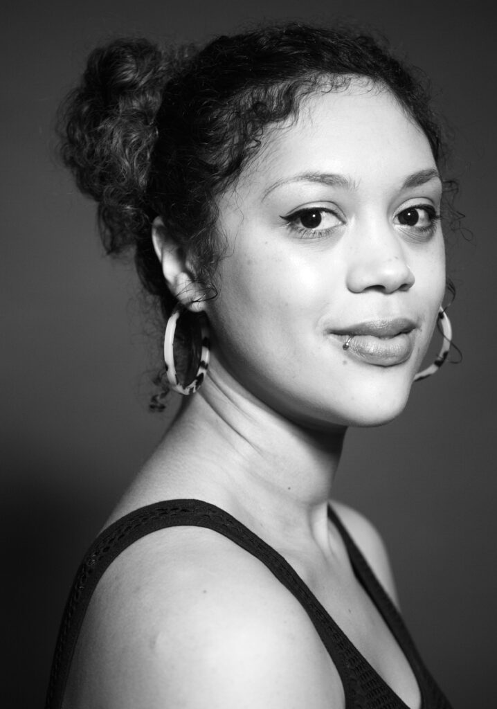 Black and white portrait of person with medium skin tone, with hoop earrings, lip piercing, curly hair in two buns, looking at the camera with slight smile.