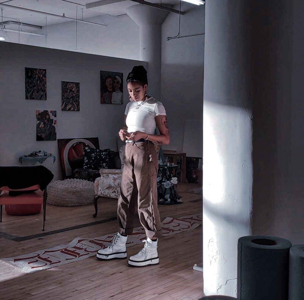 Person stands with gaze cast down in a studio space with artwork and light streaming in.