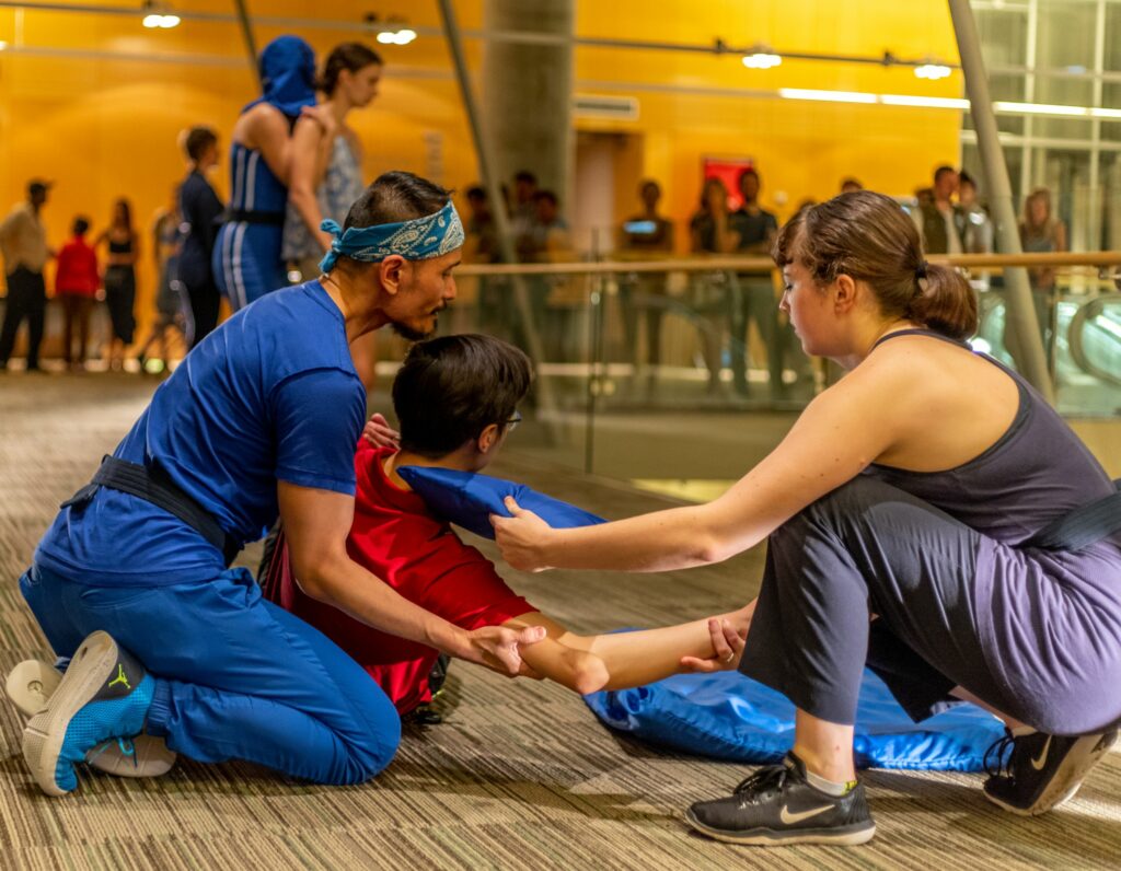 One dancer wearing a bandanna kneels holding another dancer in their arms, while a third dancer kneels and reaches out to them.