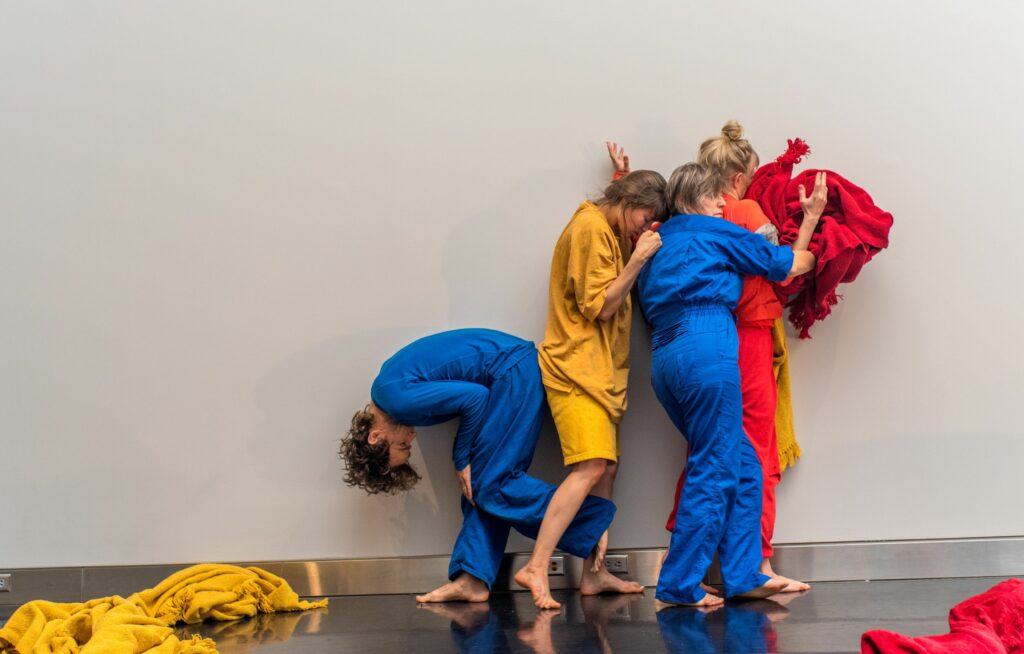 Group of dancers wearing bright blue, yellow, and red jumpsuits cluster together against white gallery wall.