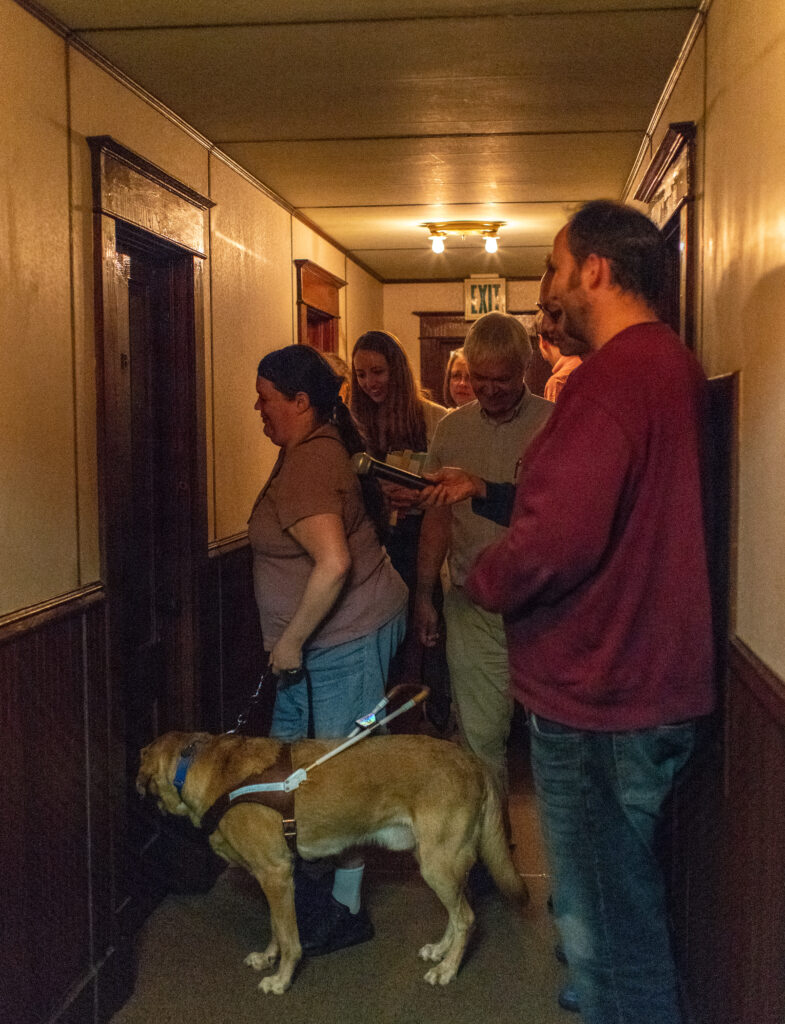 Group of people and guide dog stand in front of door in hallway.