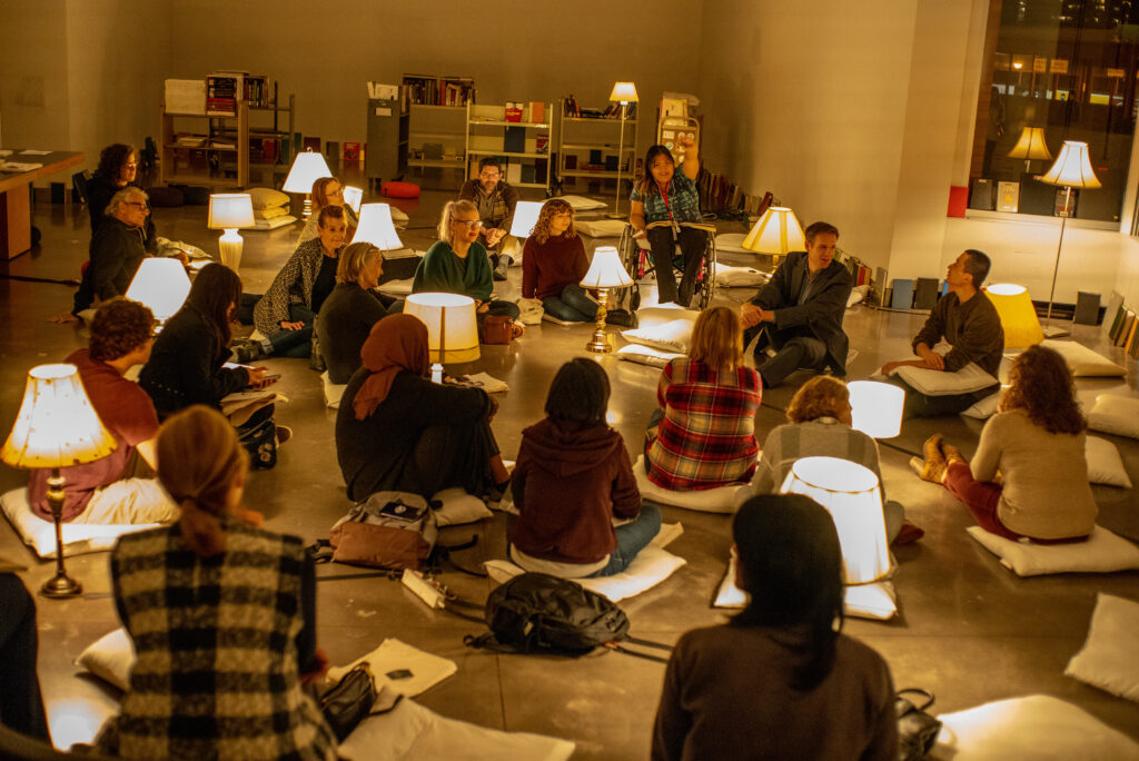 Group of people sit on floor among many lamps with shades, and bookshelves in background.