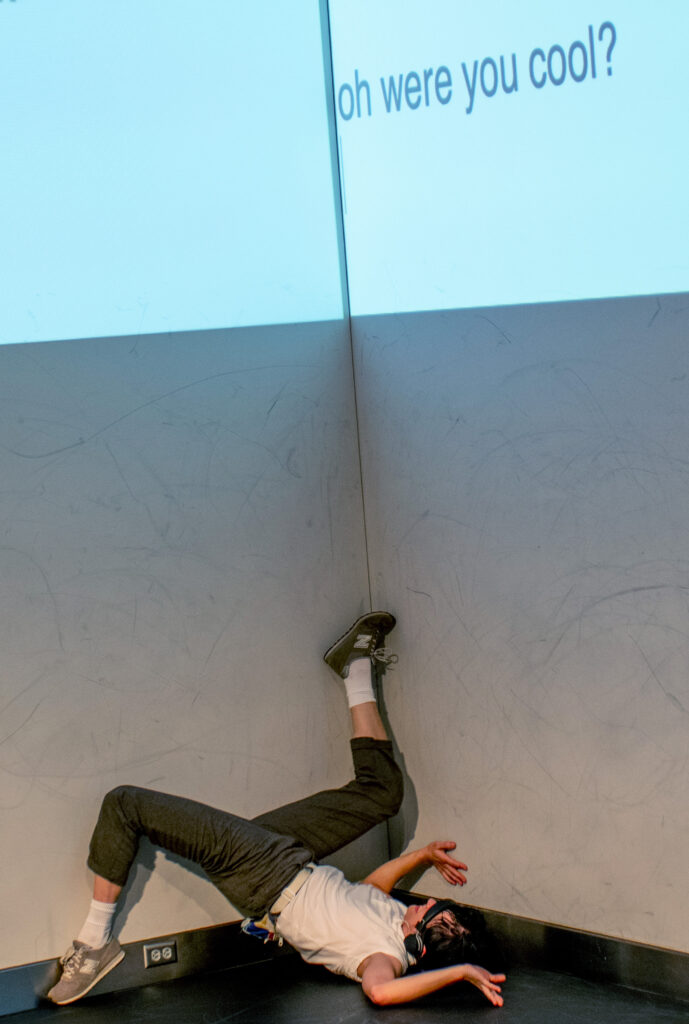 Person wearing white t-shirt and sneakers reaches their feet up into the corner of a gallery, with projection above them reading: "...were you cool?"