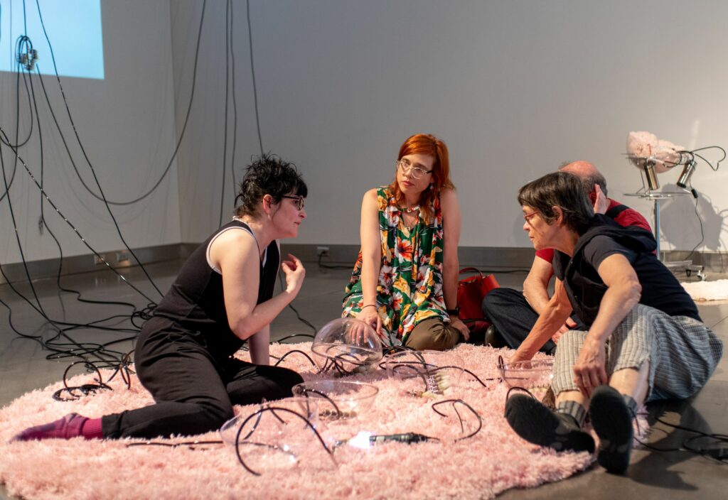 Group of people sit and talk on pink shag rug on gallery floor, with cords and lights between them.