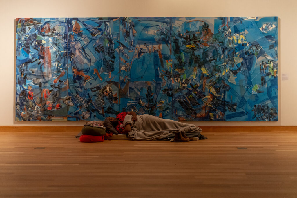 Person lays on gallery floor in front of large, blue abstract painting.