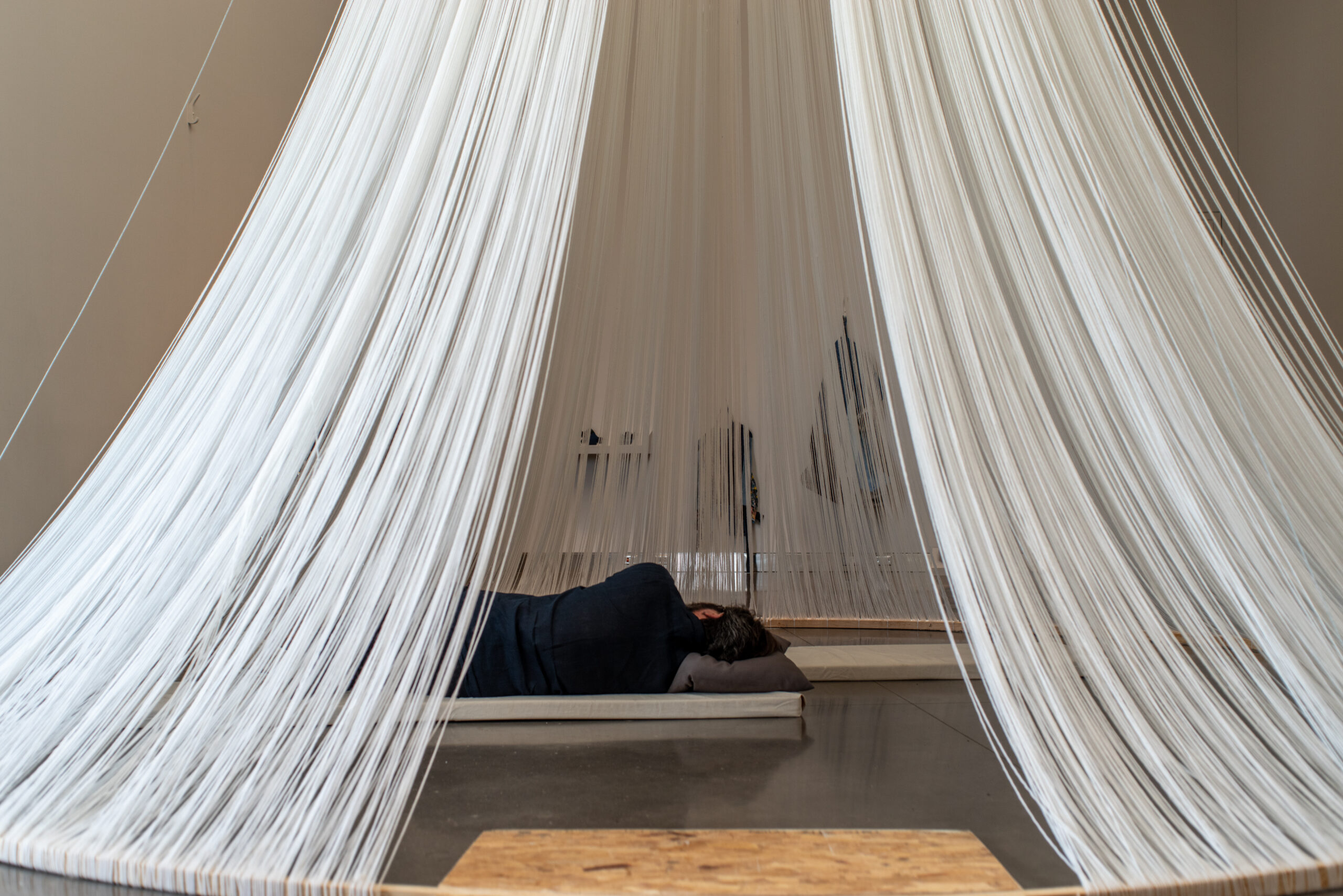 Person sleeps on floor mat with pillow, inside tent-like structure made of white string.