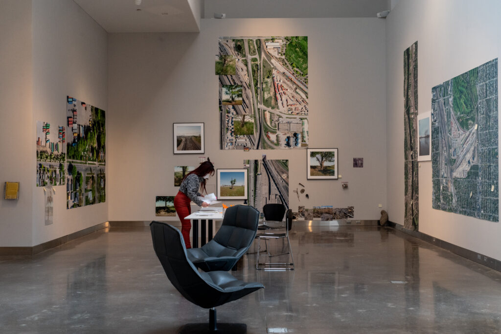 Gallery installation with many large maps and smaller framed pictures on walls, chairs on floor, and person in background.