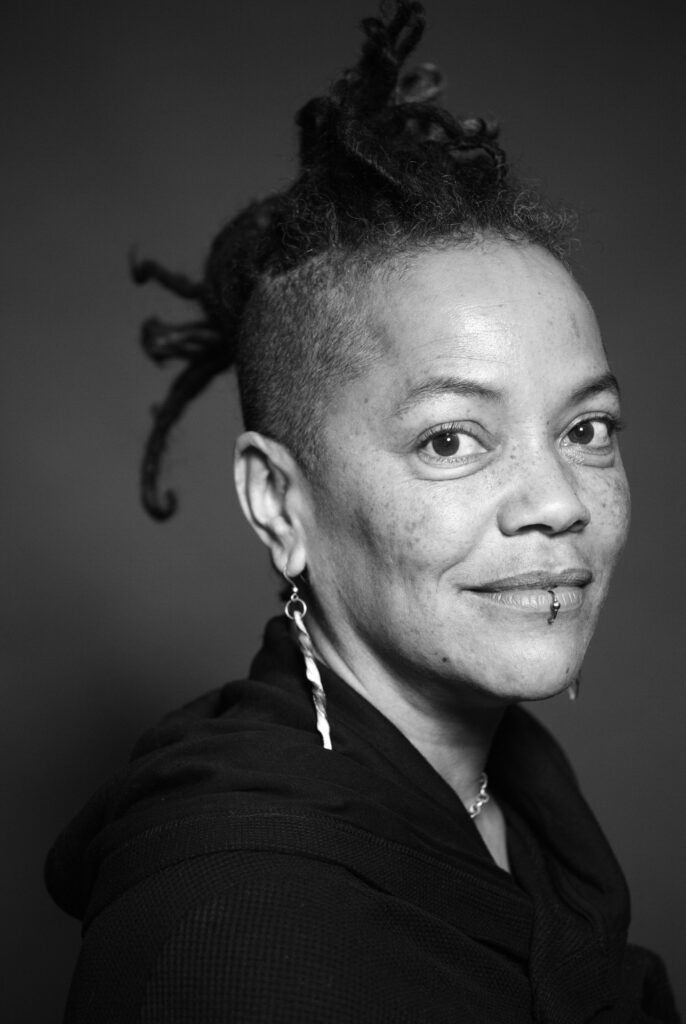 Black and white portrait of person with medium dark skin, locs and hair shaved on the sides, lip ring, long earrings, and looking at the camera with slight smile.