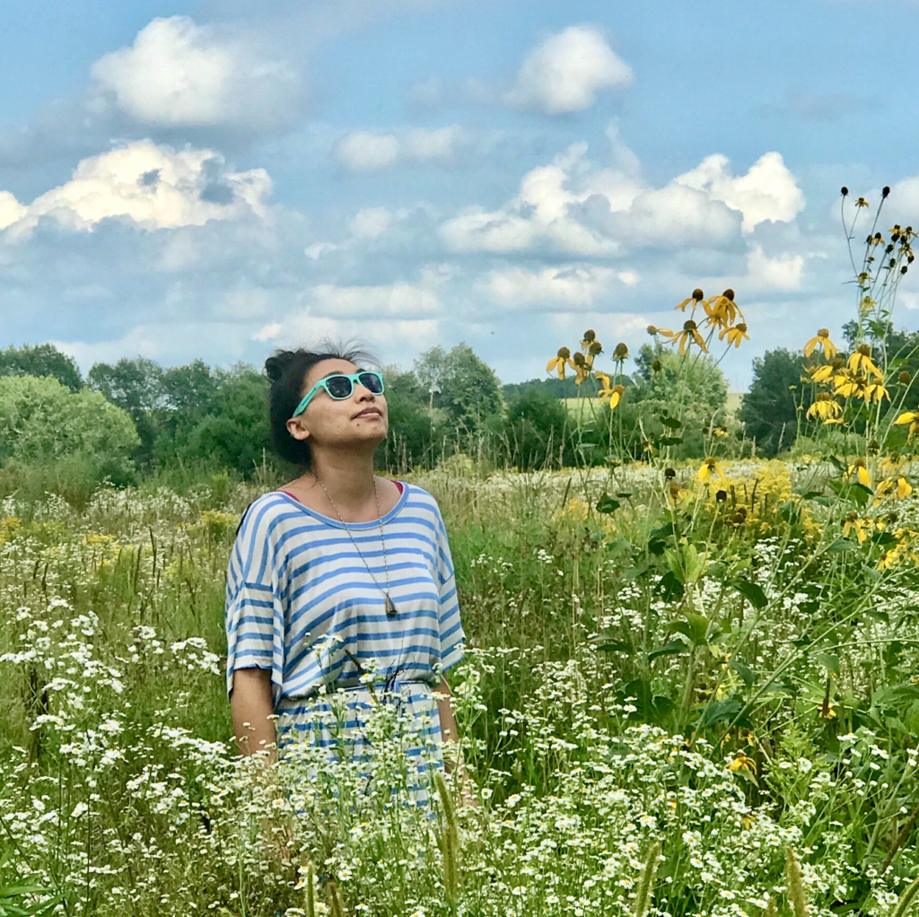 Person wearing sunglasses and blue striped t-shirt stands in flowering field looking up at blue sky.