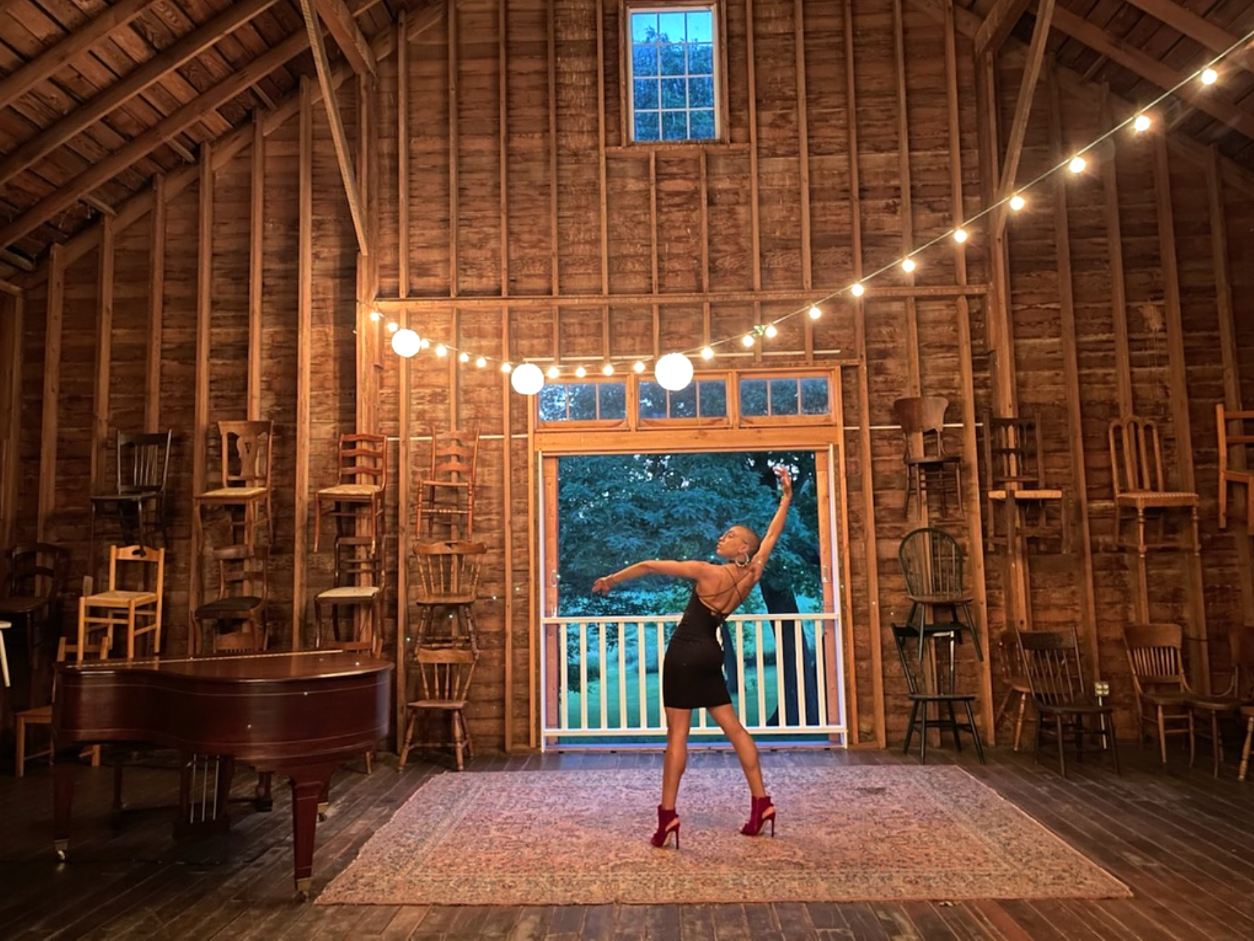 Person wearing black dress and high heels strikes pose in wooden barn with string lights, large rug, and chairs on walls.