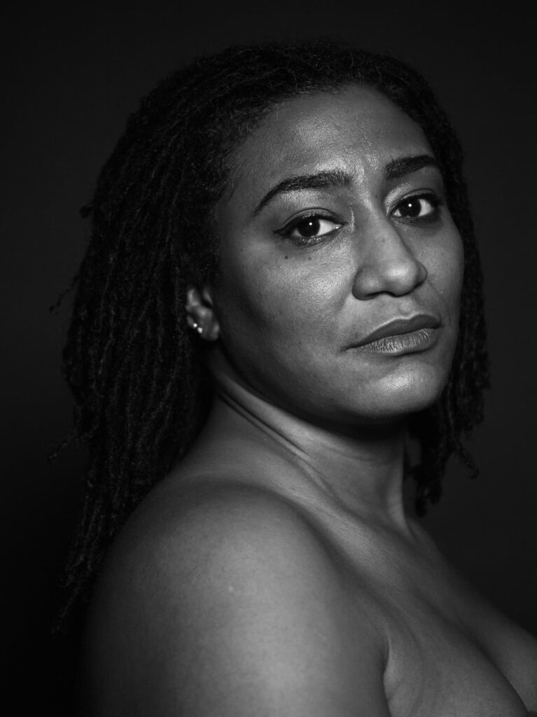 Black and white photo of person with dark skin, long hair, and exposed shoulder, head turned to camera with direct gaze.