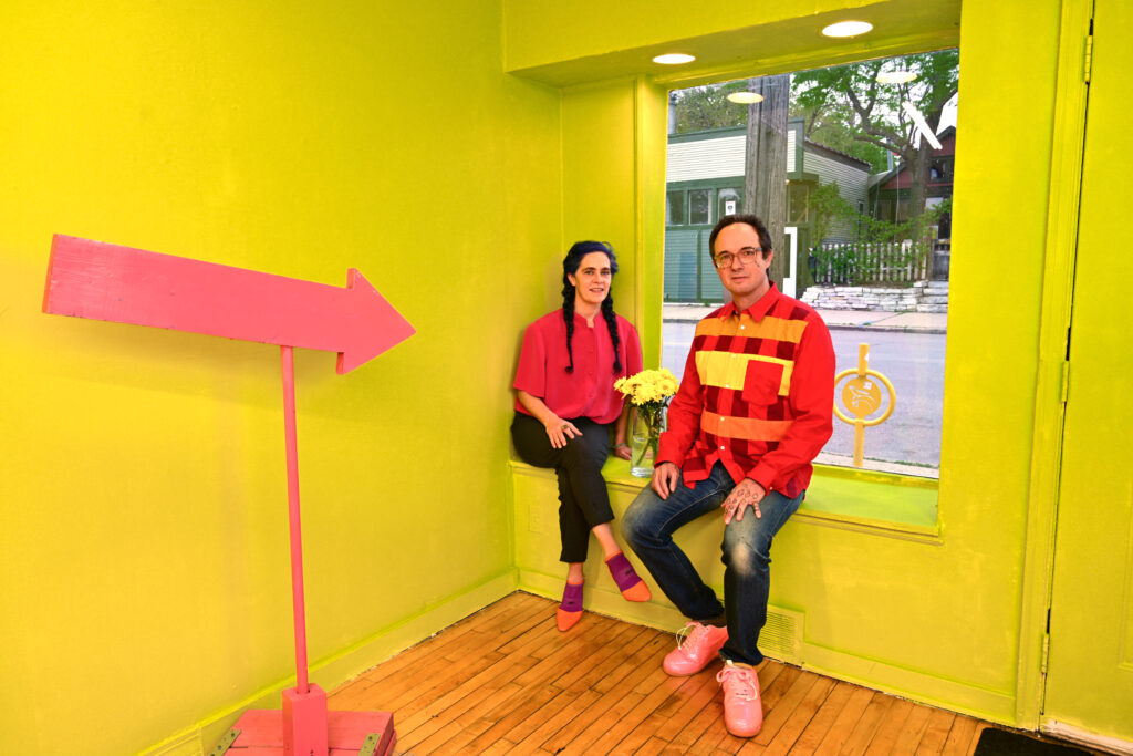 Two people wearing bright red shirts sit in window of gallery with bright chartreuse wall, and pink arrow sign pointing at them.