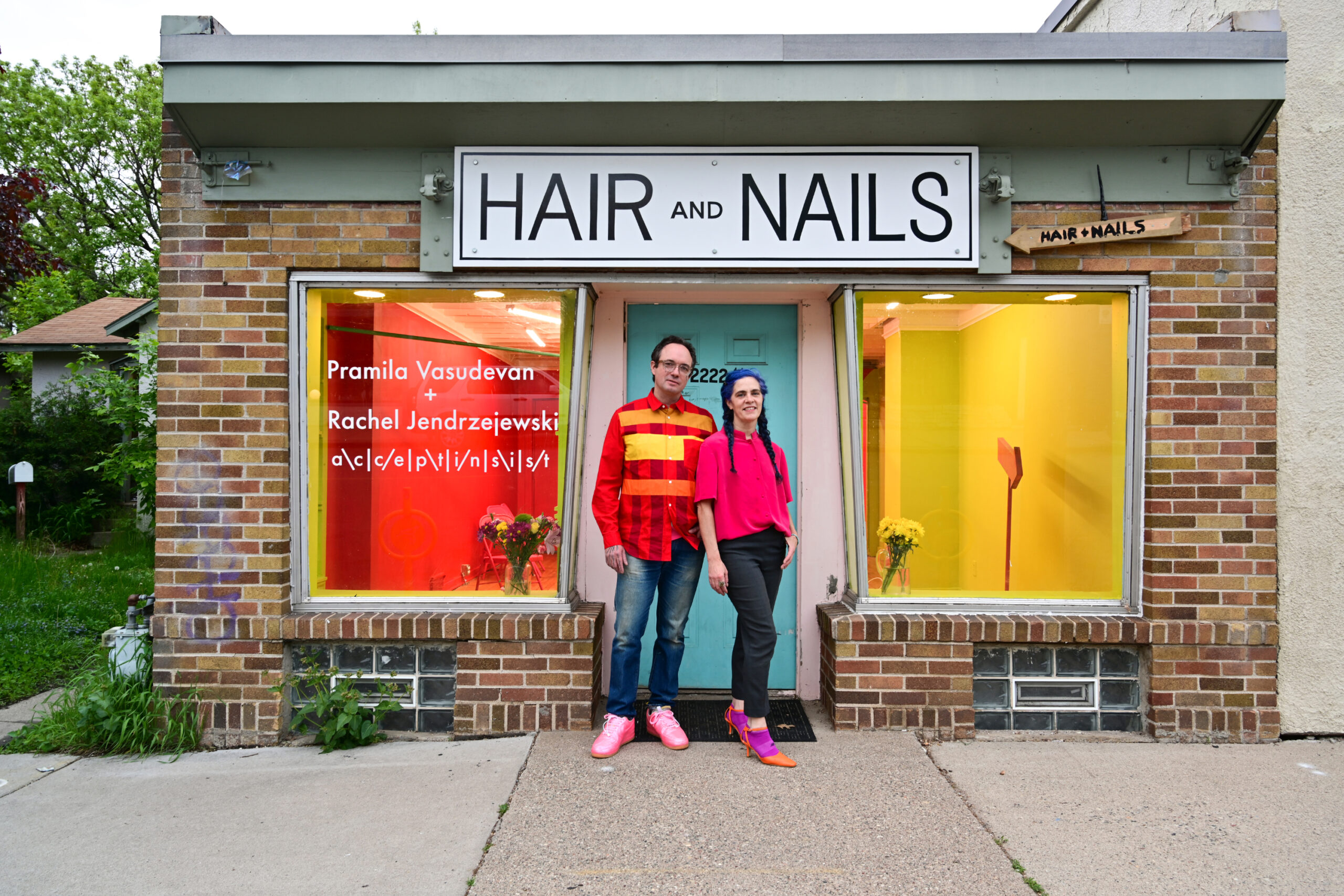 Two people stand in front of gallery with red and yellow visible through windows, teal door and sign reading HAIR and NAILS.
