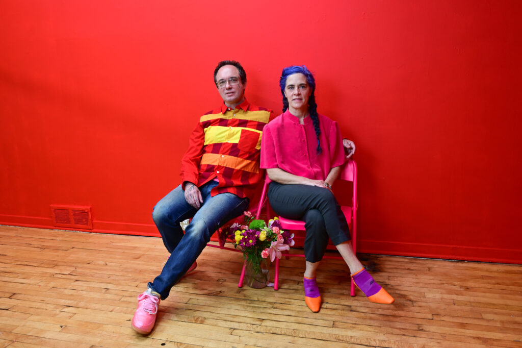 Two people wearing brightly colored shirts sit in folding chairs with legs crossed, in front of bright red wall.