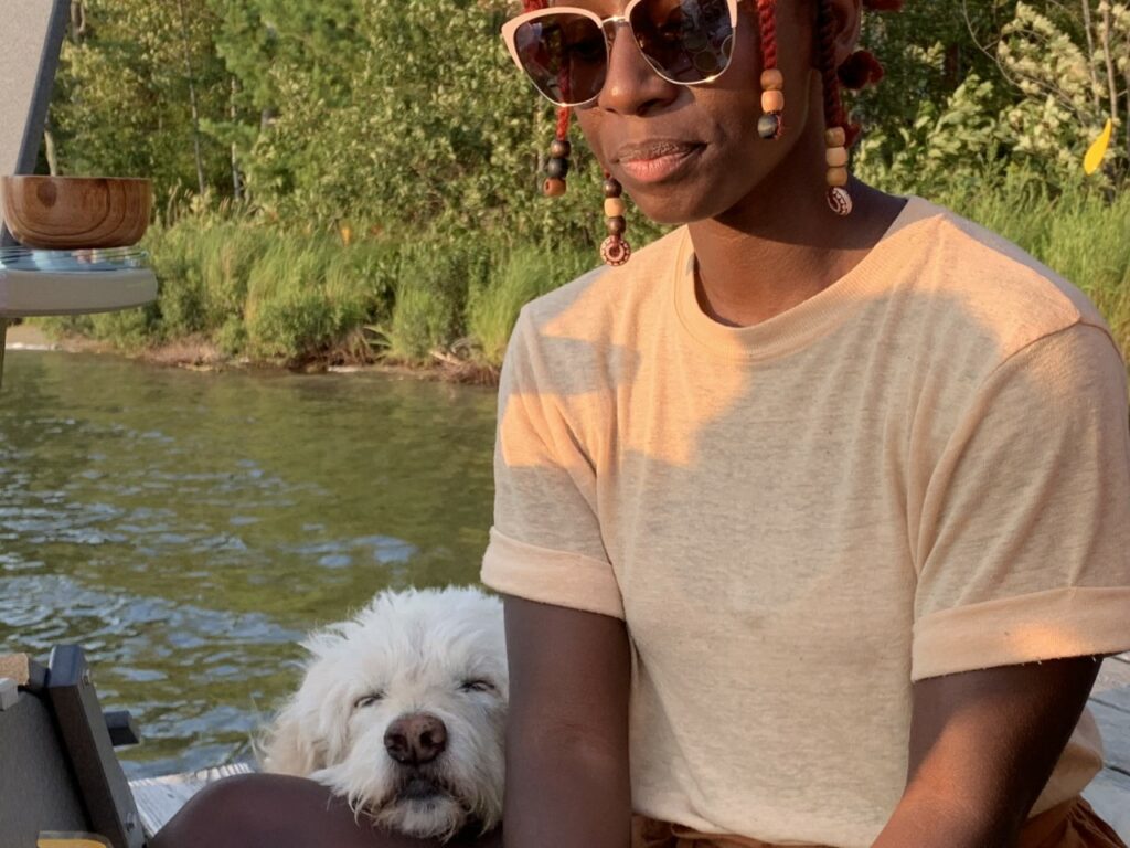 Person with dark skin and sunglasses sits by lake with dog's head resting on their thigh and foliage in background.
