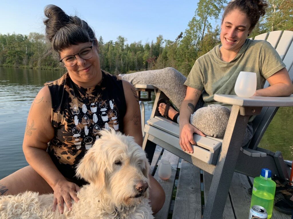 Two people smile on dock by lake, one in an Adirondack chair and the other petting a dog.